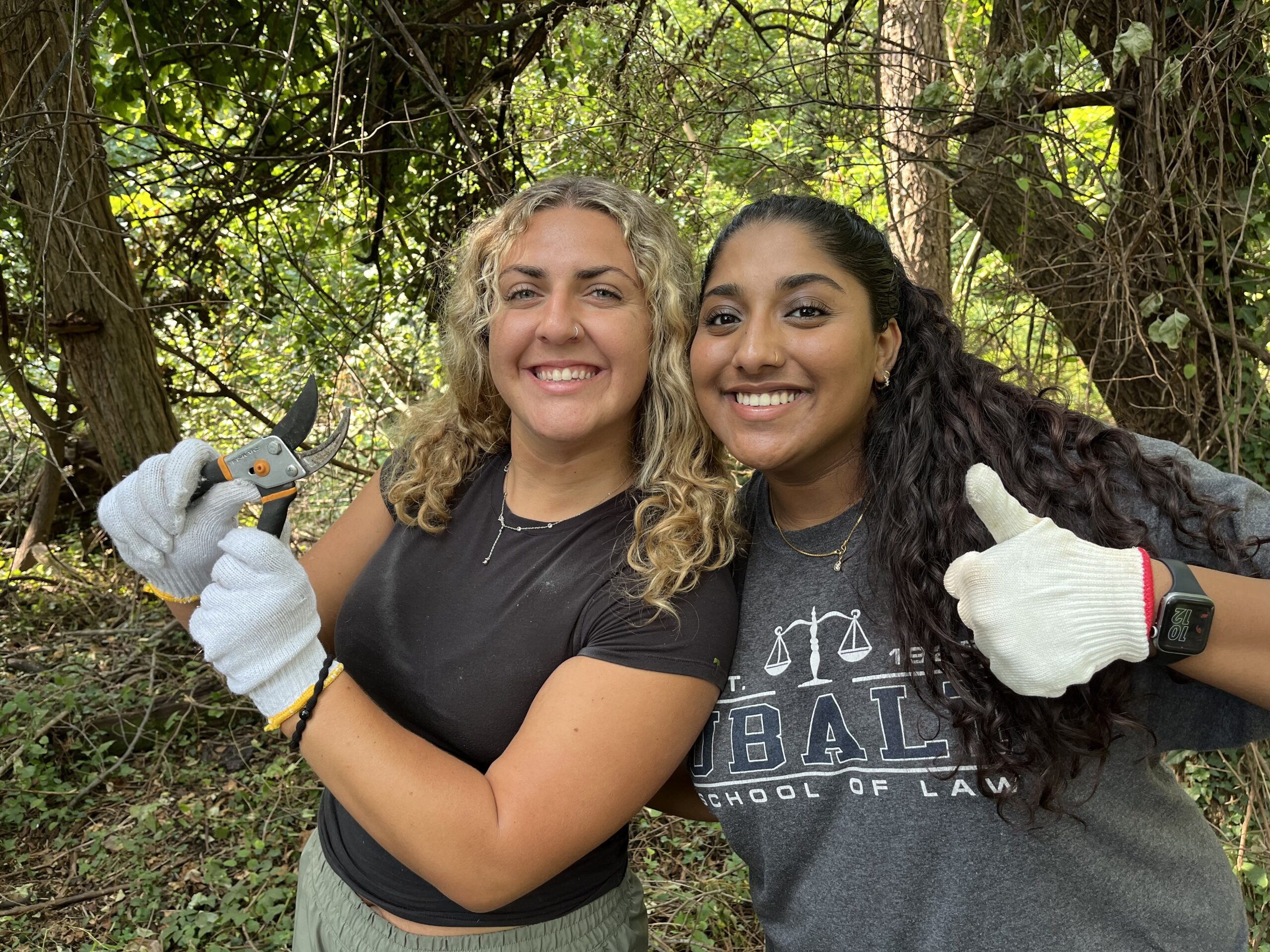 Two female students in a forest setting wearing work gloves and holding pruning shears. They are smiling and giving thumbs up.