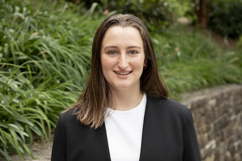 Kayla Nemirovsky in front of a stone wall, wearing a white shirt and black blazer.