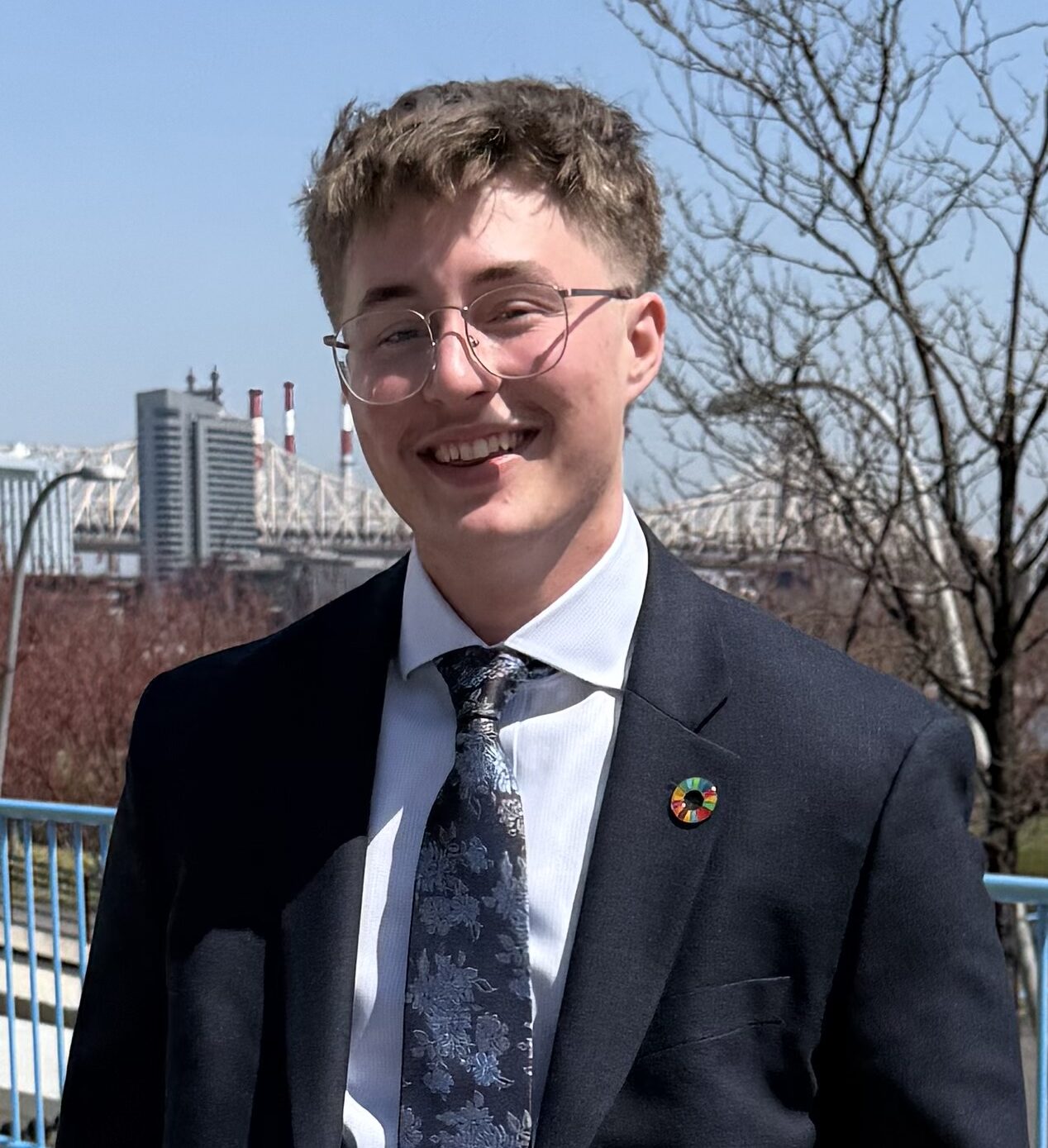 Mack Watkins in an outdoor setting with city buildings and bridge in the background, wearing a tie and navy blazer.